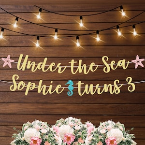 May include: Gold glitter banner with the text "Under the Sea" and "Sophie turns 3". The banner is decorated with pink and blue starfish and a blue seahorse.