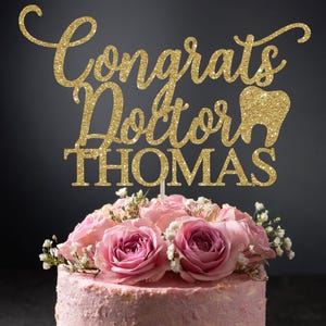 May include: Gold glitter cake topper with the text "Congrats Doctor Thomas" and a tooth graphic. The topper is on a pink cake with pink roses.