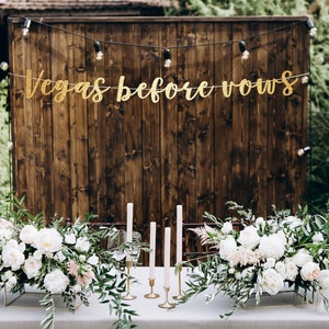 May include: A wedding table setting with a rustic wooden backdrop and a gold glitter banner that reads "Vegas before vows". The table is decorated with white flowers and greenery, and there are candles in gold holders.