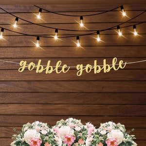 K&ouml;nnte beinhalten: Ein goldener Glitzerbanner mit dem Text "gobble gobble" h&auml;ngt an einer Holzwand mit Lichterketten. Der Banner ist perfekt f&uuml;r eine Thanksgiving-Party oder ein Treffen.