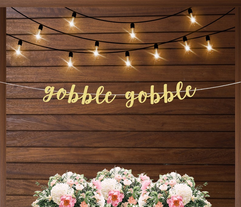 K&ouml;nnte beinhalten: Ein goldener Glitzerbanner mit dem Text "gobble gobble" h&auml;ngt an einer Holzwand mit Lichterketten. Der Banner ist perfekt f&uuml;r eine Thanksgiving-Party oder ein Treffen.