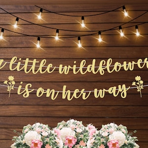 May include: A gold glitter banner with the text "a little wildflower is on her way" and two small flower accents on either side. The banner is hanging on a rustic wood background with string lights.