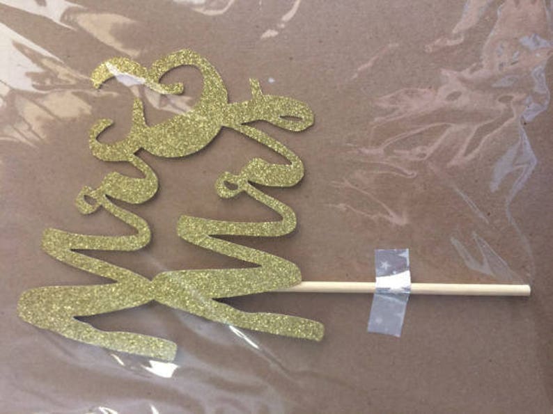Puede incluir: Topper para pastel de brillo dorado con las palabras "Mr & Mrs" en un palo de madera.