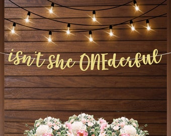 May include: Gold glitter banner with the text "isn't she ONEderful" hanging on a wood wall with string lights.