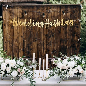 May include: A wedding reception table setting with white flowers, greenery, and candles in front of a wooden wall with a gold glitter banner that reads "#WeddingHashtag".