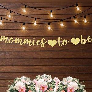 May include: Gold glitter banner with the text "Mommies to be" and two gold glitter hearts. The banner is hanging on a wood wall with string lights.