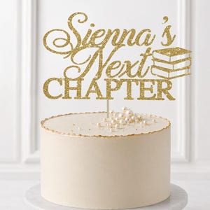 May include: A gold glitter cake topper featuring the text Sienna's Next CHAPTER with a small stack of books icon. The topper is mounted on a thin stick and is positioned above a round white cake decorated with small white pearls on top.