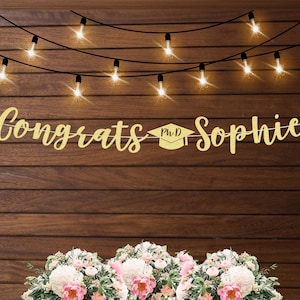 May include: Gold glitter banner with the text "Congrats PhD Sophie" hanging on a wood wall with string lights. The banner is decorated with a graduation cap.