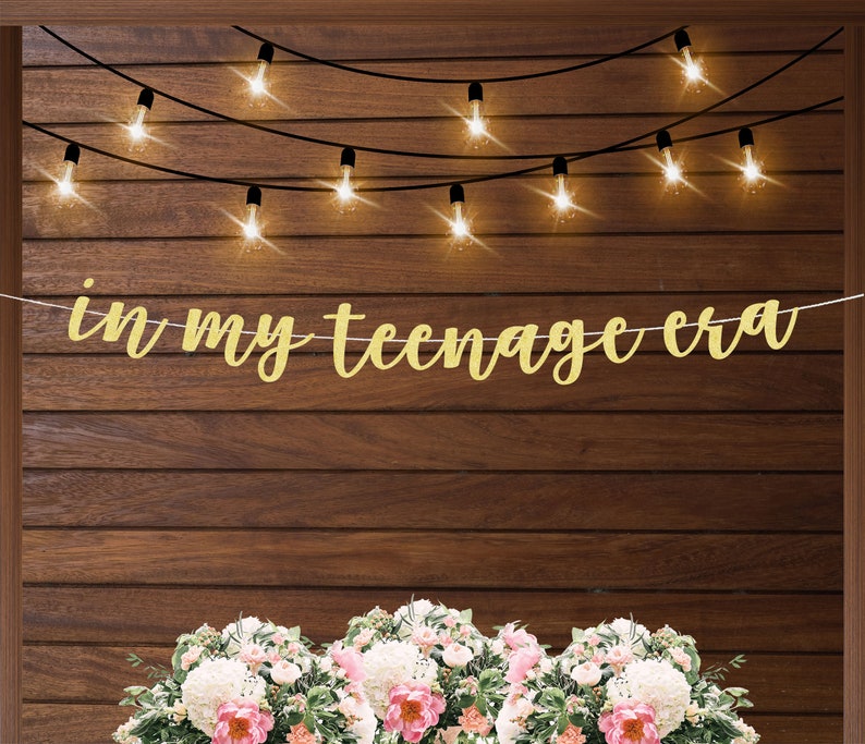 May include: A gold glitter banner with the text 'in my teenage era' hanging on a brown wood wall with string lights. The banner is hanging in front of three bouquets of white and pink flowers.