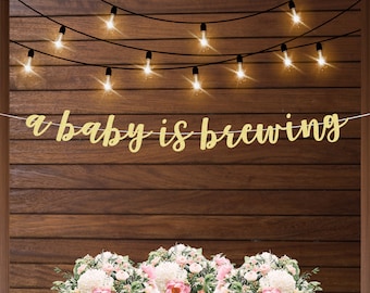 May include: A gold glitter banner with the text "a baby is brewing" hanging on a wood wall with string lights. The banner is decorated with flowers.
