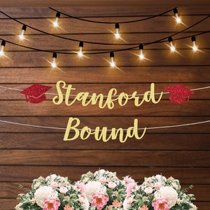 May include: Gold glitter banner with the text "Stanford Bound" and red glitter graduation caps on each end. The banner is hanging on a wood wall with string lights.