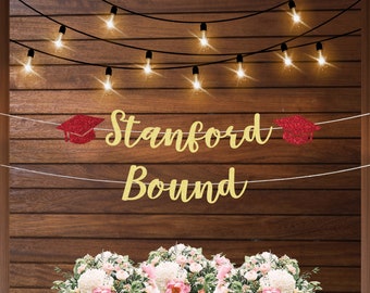 May include: Gold glitter banner with the text "Stanford Bound" and red glitter graduation caps on each end. The banner is hanging on a wood wall with string lights.