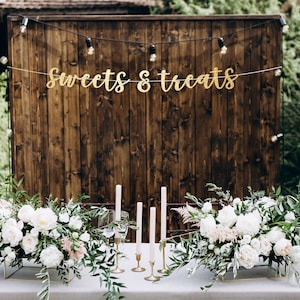 May include: A rustic wooden backdrop with a gold banner that reads "Sweets & Treats". Two white floral arrangements with greenery are on a table in front of the backdrop. There are candles and glasses on the table.