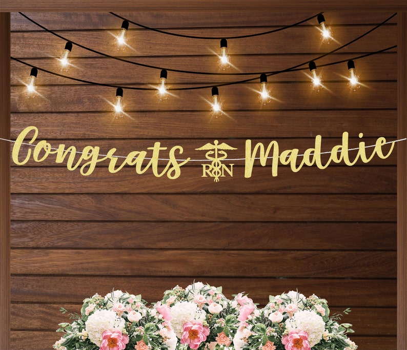 May include: Gold glitter banner with the text "Congrats Maddie" and a medical symbol with the letters "RN" in the center. The banner is hanging against a wood background with string lights.