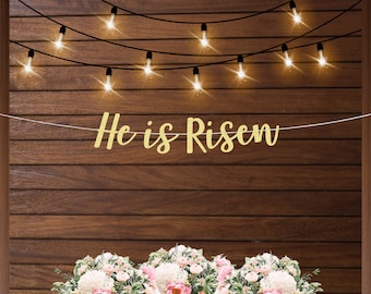 May include: Gold glitter banner with the text "He is Risen" hanging on a wood wall with string lights. The banner is in front of a floral arrangement with white and pink flowers.