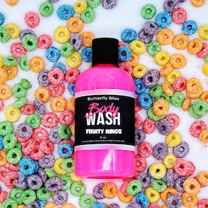 Puede incluir: Botella rosa de gel de baño con una etiqueta negra que dice "Butterfly Bliss Body Wash Fruity Rings 8 oz sustainably sourced | made in the USA". La botella está rodeada de anillos de cereales coloridos.