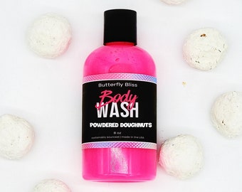 May include: Pink body wash bottle with black lettering that reads "Butterfly Bliss Body Wash Powdered Doughnuts 8 oz sustainably sourced | made in the USA". The bottle is surrounded by white bath bombs.