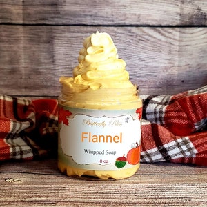 Pode incluir: Um frasco de vidro de sabonete batido com um redemoinho amarelo e laranja no topo. O frasco tem um rótulo que diz "Butterfly Bliss" e "Flannel Whipped Soap 8 oz".