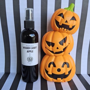 May include: A black bottle of body spray with a white label that says "Butterfly Kisses Wicked Candy Apple Body Spray 4 oz". The bottle is in front of a stack of three orange pumpkin-shaped decorations with black jack-o-lantern faces.
