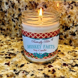 May include: A clear glass jar candle with a red, white, and blue label that says "Butterfly Bliss Monkey Farts Soy Candle 9oz".