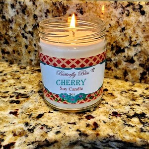 May include: A lit soy candle in a glass jar with a red, white, and blue checkered label. The label reads "Butterfly Bliss Cherry Soy Candle 9oz".