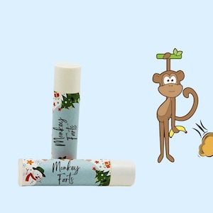 Puede incluir: Dos tubos de bálsamo labial con una etiqueta azul y blanca que dice "Monkey Farts". La etiqueta tiene un muñeco de nieve y un árbol de Navidad. Los tubos de bálsamo labial están sobre un fondo azul claro.