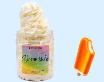May include: A jar of whipped body butter with a label that reads "Butterfly Bliss Dreamsicle Whipped Body Butter 8 oz". The jar is filled with a white whipped cream-like product. A small orange popsicle with a bite taken out of it is in the background.