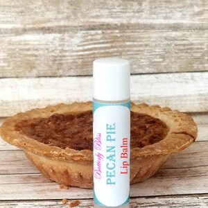May include: A white tube of lip balm with a blue and pink label that says "Butterfly Bliss Pecan Pie Lip Balm". The lip balm is sitting on a wooden surface next to a pecan pie.
