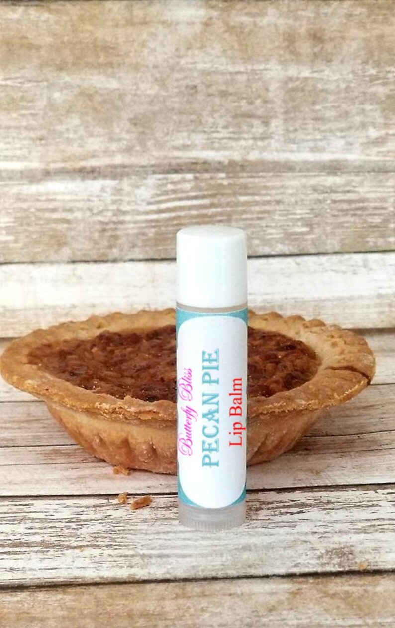 May include: A white tube of lip balm with a blue and pink label that says "Butterfly Bliss Pecan Pie Lip Balm". The lip balm is sitting on a wooden surface next to a pecan pie.