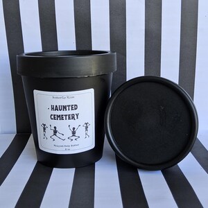 K&ouml;nnte beinhalten: Schwarzes Glas mit geschlagenem K&ouml;rperbutter mit einem wei&szlig;en Etikett, auf dem "Haunted Cemetery" steht und vier Skelette abgebildet sind. Das Glas steht auf einer schwarz-wei&szlig; gestreiften Oberfl&auml;che.