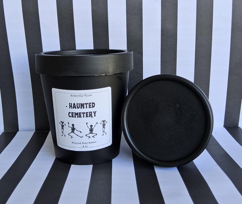 K&ouml;nnte beinhalten: Schwarzes Glas mit geschlagenem K&ouml;rperbutter mit einem wei&szlig;en Etikett, auf dem "Haunted Cemetery" steht und vier Skelette abgebildet sind. Das Glas steht auf einer schwarz-wei&szlig; gestreiften Oberfl&auml;che.