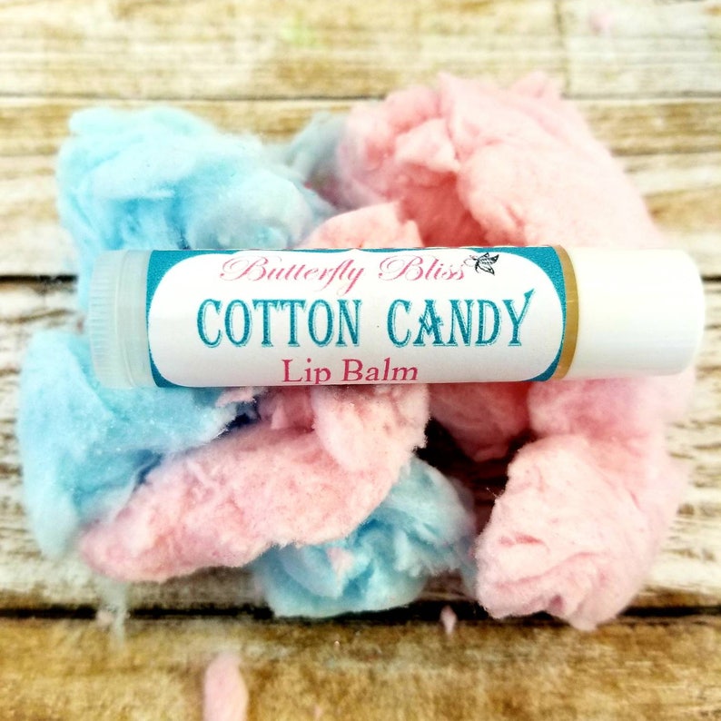 May include: A white lip balm tube with a blue and gold label that reads "Butterfly Bliss Cotton Candy Lip Balm". The tube is surrounded by pink and blue cotton candy.