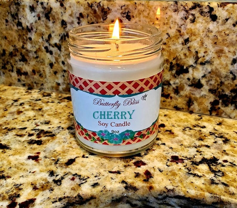 May include: A lit soy candle in a glass jar with a red, white, and blue checkered label. The label reads "Butterfly Bliss Cherry Soy Candle 9oz".