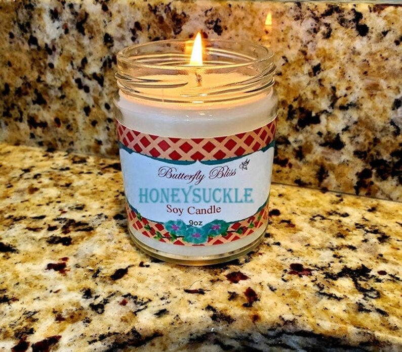 K&ouml;nnte beinhalten: Eine brennende Sojawachskerze in einem durchsichtigen Glasgef&auml;&szlig; mit einem rot-wei&szlig;-blauen Etikett, auf dem "Butterfly Bliss Honeysuckle Soy Candle 9oz" steht.