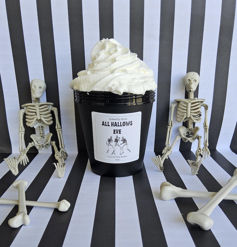 K&ouml;nnte beinhalten: Ein schwarzer Beh&auml;lter mit einem wei&szlig;en Etikett, auf dem "Butterfly Kisses All Hallows Eve Whipped Body Butter 8 oz" steht, ist von zwei wei&szlig;en Skeletten aus Plastik und zwei wei&szlig;en Knochen aus Plastik umgeben. Der Beh&auml;lter ist mit wei&szlig;er, geschlagenem K&ouml;rperbutter gef&uuml;llt.
