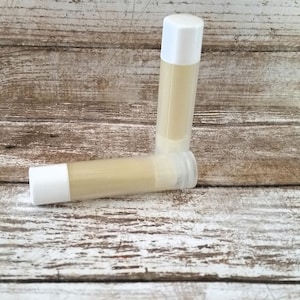 May include: Two white tubes of lip balm, one lying on its side and the other standing upright. The lip balm is a light yellow color.