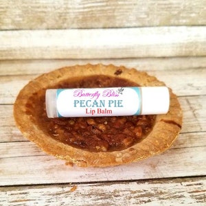 May include: A white lip balm tube with a blue and pink label that says "Butterfly Bliss Pecan Pie Lip Balm". The lip balm is sitting on top of a mini pecan pie.