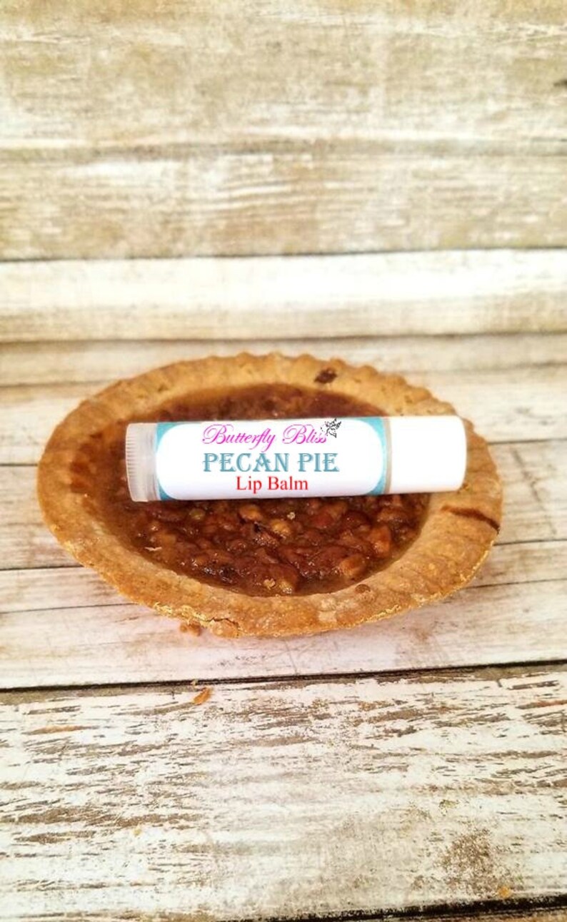 May include: A white lip balm tube with a blue and pink label that says "Butterfly Bliss Pecan Pie Lip Balm". The lip balm is sitting on top of a mini pecan pie.