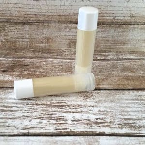 May include: Two white tubes of lip balm on a wooden surface. The lip balm is a light yellow color.