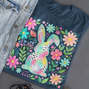Könnte beinhalten: Marineblaues T-Shirt mit einem bunten Hasen-Design, umgeben von Blumen. Der Hase ist mit Pastellfarben und schwarzen Punkten bemalt. Rosa, blaue und pfirsichfarbene Blumen sind um den Hasen angeordnet. Eine Jeansjacke, Armbänder und Sonnenbrillen sind ebenfalls im Bild.