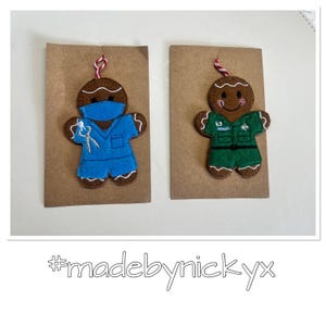 May include: Two felt gingerbread ornaments attached to brown cardstock. One gingerbread figure wears a blue scrub top and mask with a dental tool graphic. The other wears a green uniform with a belt. Both have red and white hanging strings.