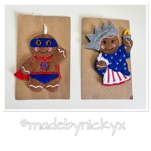 May include: Two felt gingerbread ornaments on brown cardstock. One is a superhero with a blue mask, cape, and a letter G on its chest. The other is dressed as the Statue of Liberty with a crown labelled Liberty and a torch. Text reads #madebynickyx.