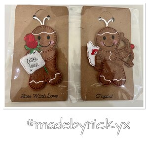 May include: Two brown felt gingerbread person ornaments on cardboard backing. One holds a red rose and a white tag reading With love. The other is dressed as Cupid with white wings and a brown bow. Both have white stitching and pink cheeks.