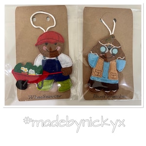 May include: Two felt gingerbread ornaments on cardboard backing. One depicts a gardener in a red cap, white shirt, blue overalls, and green boots pushing a red wheelbarrow with plants. The other is a bird watcher with glasses and a vest.