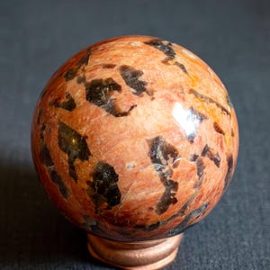 Könnte beinhalten: Ein polierter, kugelförmiger Stein mit glatter, glänzender Oberfläche, der ein gesprenkeltes Muster in warmen Orange-, Pfirsich- und Beigetönen aufweist, durchsetzt mit unregelmäßigen, dunkelbraunen bis schwarzen Einschlüssen auf einem kupferfarbenen Sockel.