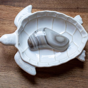 May include: A white ceramic turtle-shaped dish with a gray and white stone inside. The dish has a textured surface and is placed on a wooden table. The stone has a smooth, polished appearance.