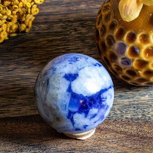 Puede incluir: Una piedra esférica pulida que presenta un patrón jaspeado de inclusiones minerales en azul profundo y blanco. El objeto liso y redondeado muestra una textura superficial brillante con variaciones de color distintas en toda su forma compacta.