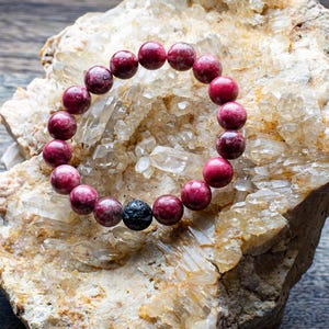 Peut inclure: Un bracelet en pierres précieuses roses avec une perle de lave noire. Le bracelet est posé sur un lit de cristaux de quartz blancs.