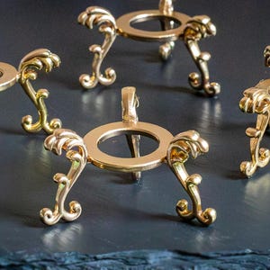 Puede incluir: Varios soportes de metal dorado ornamentados, con una parte superior circular y patas curvas decorativas. Los soportes están dispuestos sobre una superficie oscura y texturizada, mostrando su intrincado diseño y su uso potencial como expositores.