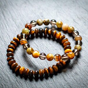 Pode incluir: Duas pulseiras de contas com uma mistura de contas castanhas, laranja e transparentes. As pulseiras estão sobrepostas, mostrando as diferentes texturas e cores das pedras. As pulseiras estão sobre uma superfície cinza escuro.